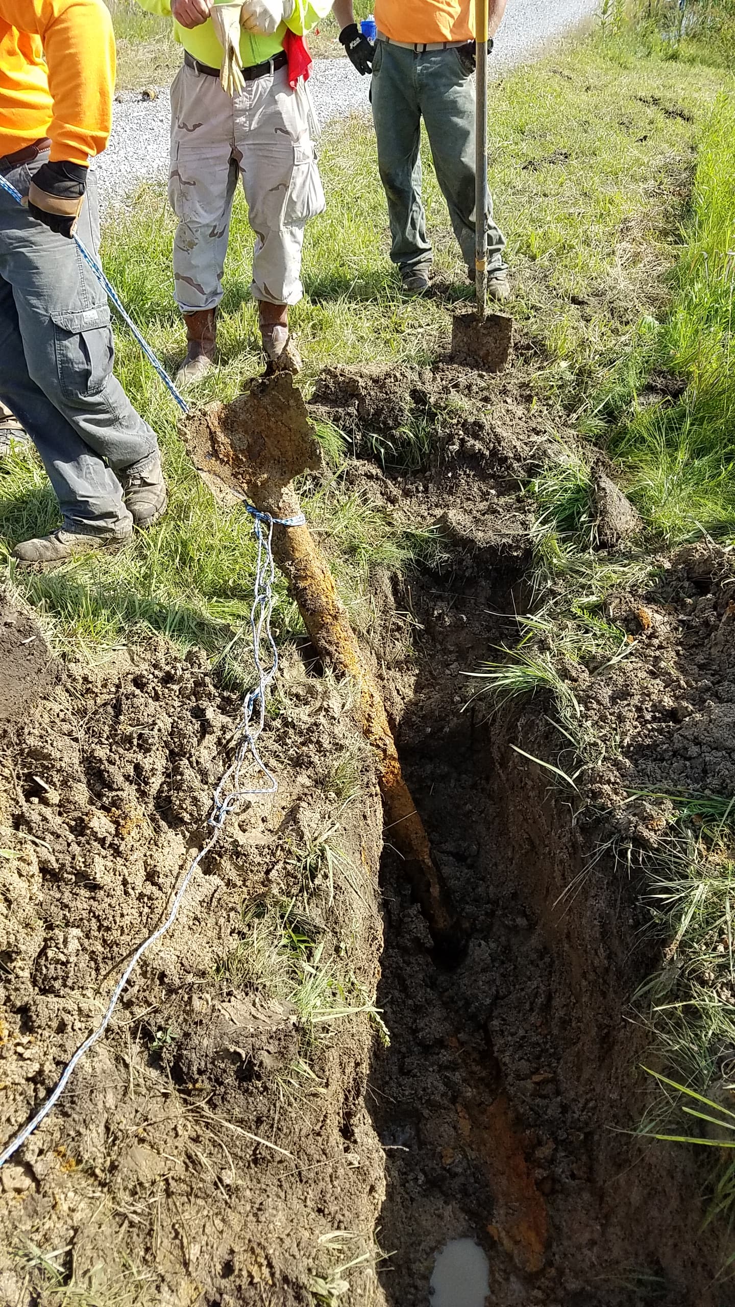 Workers use a rope to pull a large, rusty, finned metal object from a narrow trench.
