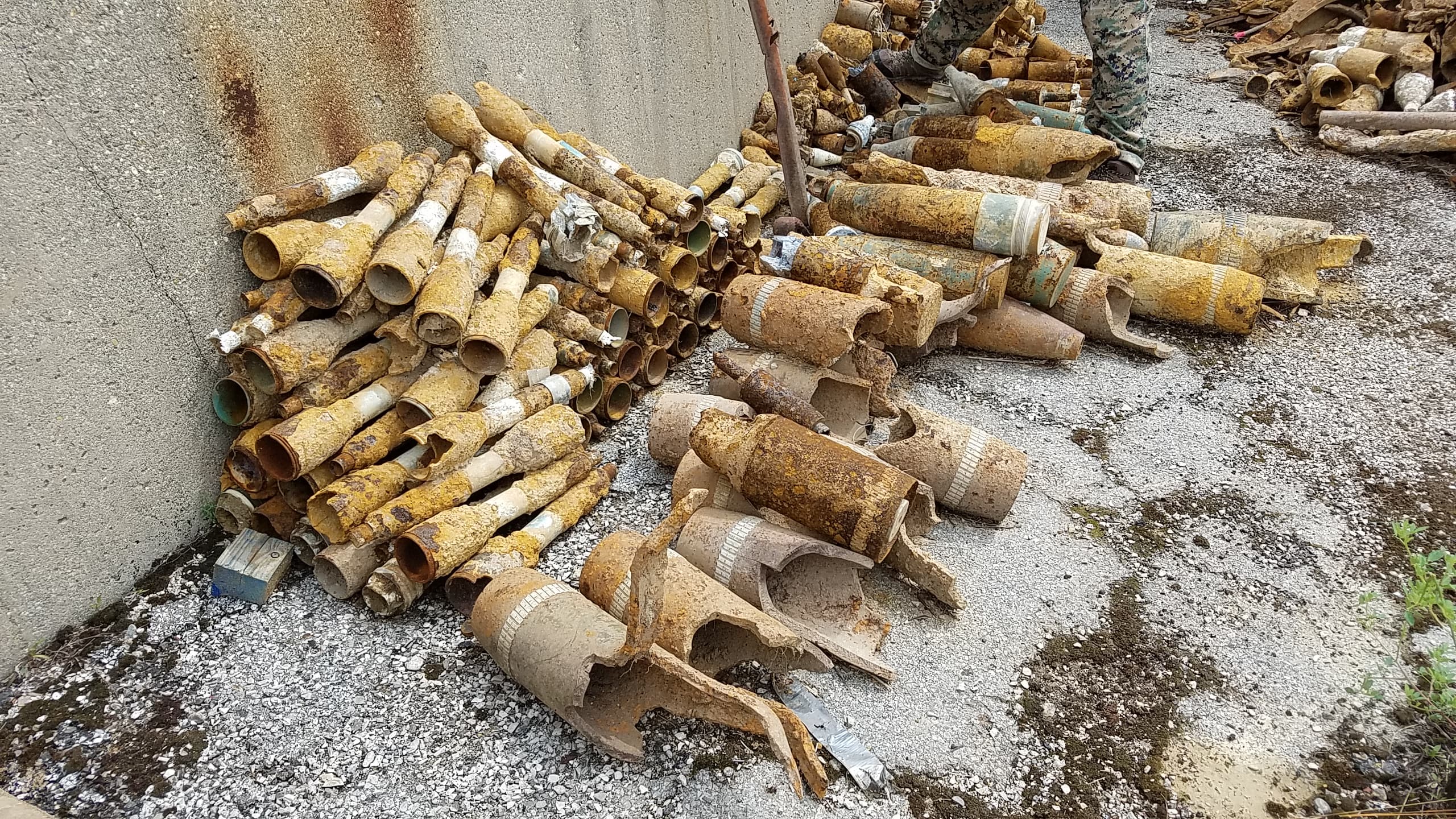Numerous rusted, decaying mortar rounds and artillery shells stacked against a weathered concrete wall.